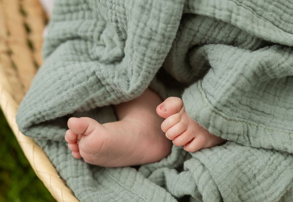 Tiny baby feet peeking out from sage green muslin wrap in woven basket.
