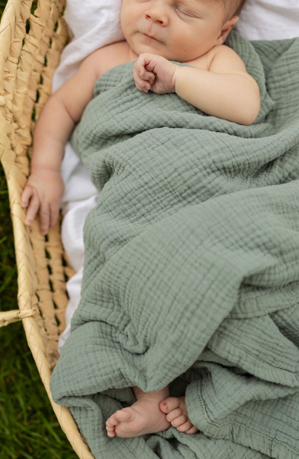 Sleeping newborn wrapped in sage green muslin blanket in natural woven basket.