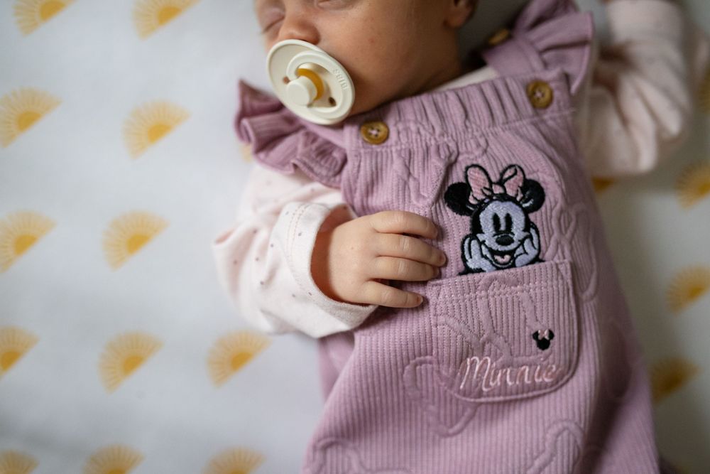 Baby wearing pink knitted sweater with Mickey Mouse design rests against yellow star patterned background.