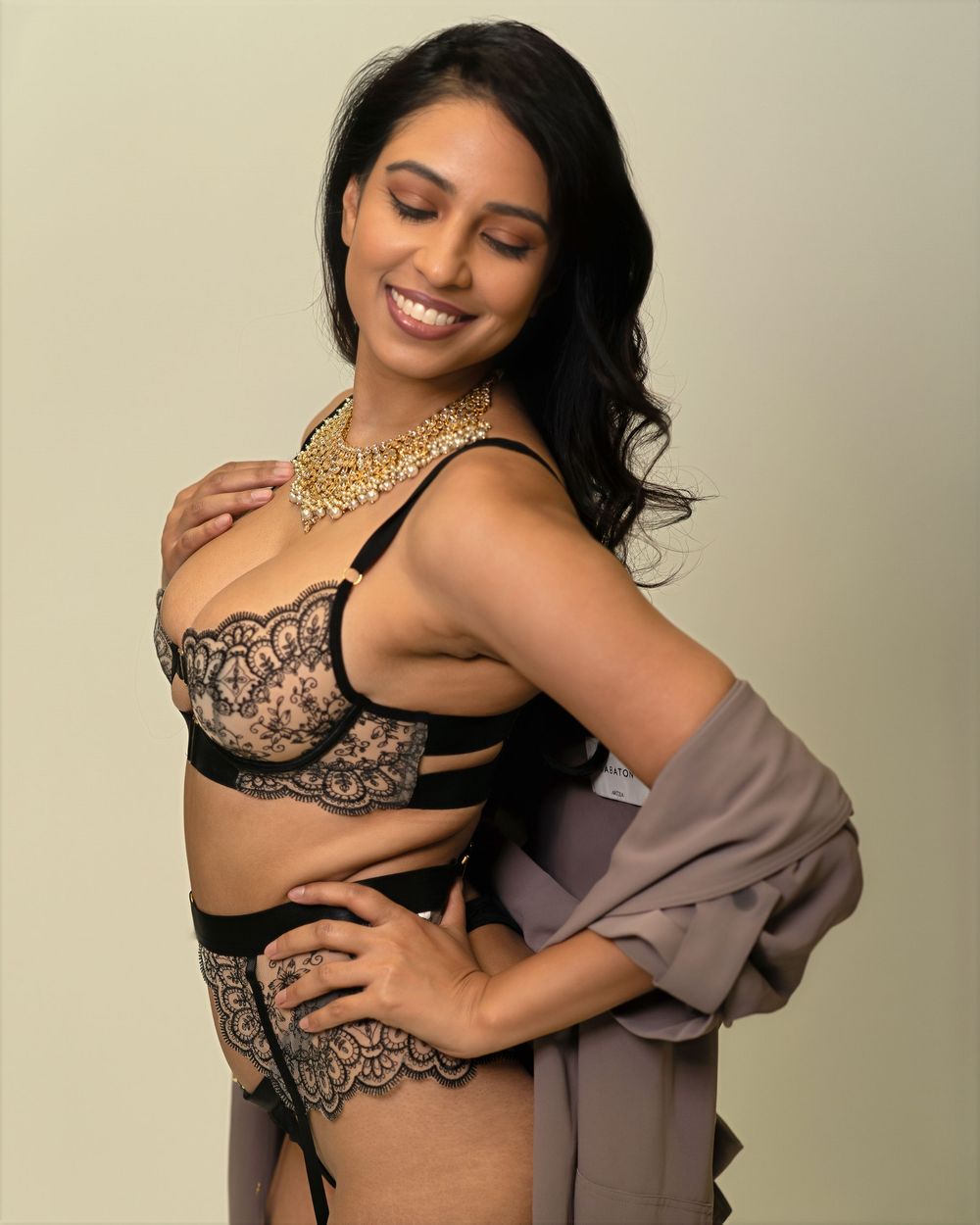 Woman smiling in black lace lingerie set with gold necklace and gray robe, posing against neutral background.