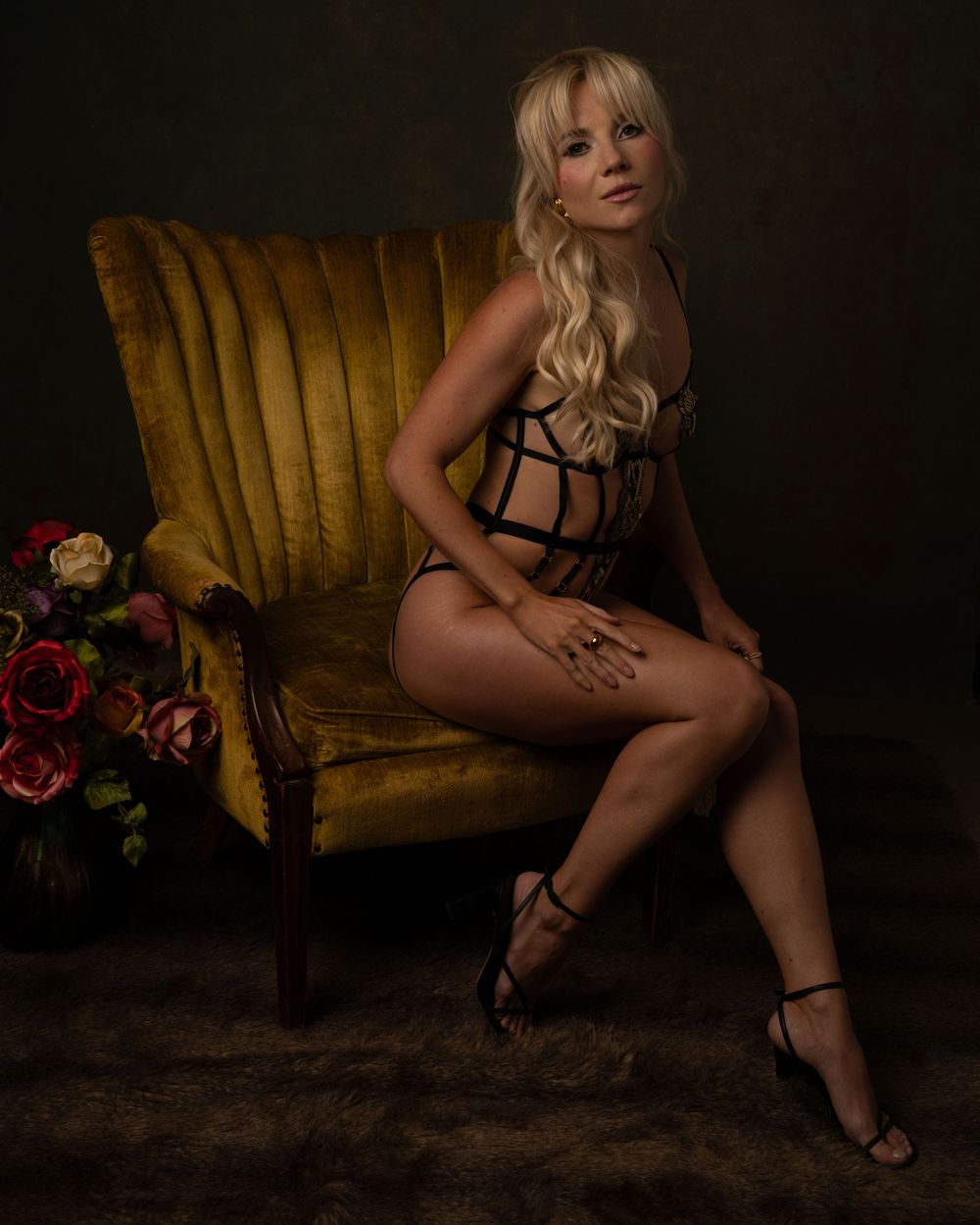 Blonde woman in black strappy lingerie sits on gold velvet chair in dramatic studio portrait.
