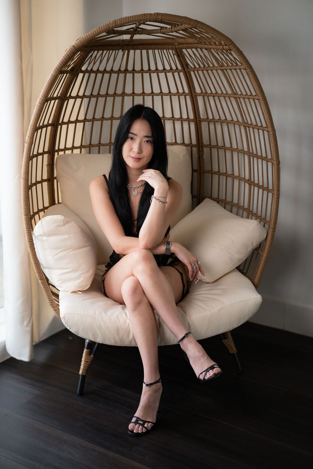 A dark-haired woman in a black dress sits elegantly in a wicker egg chair by a window.