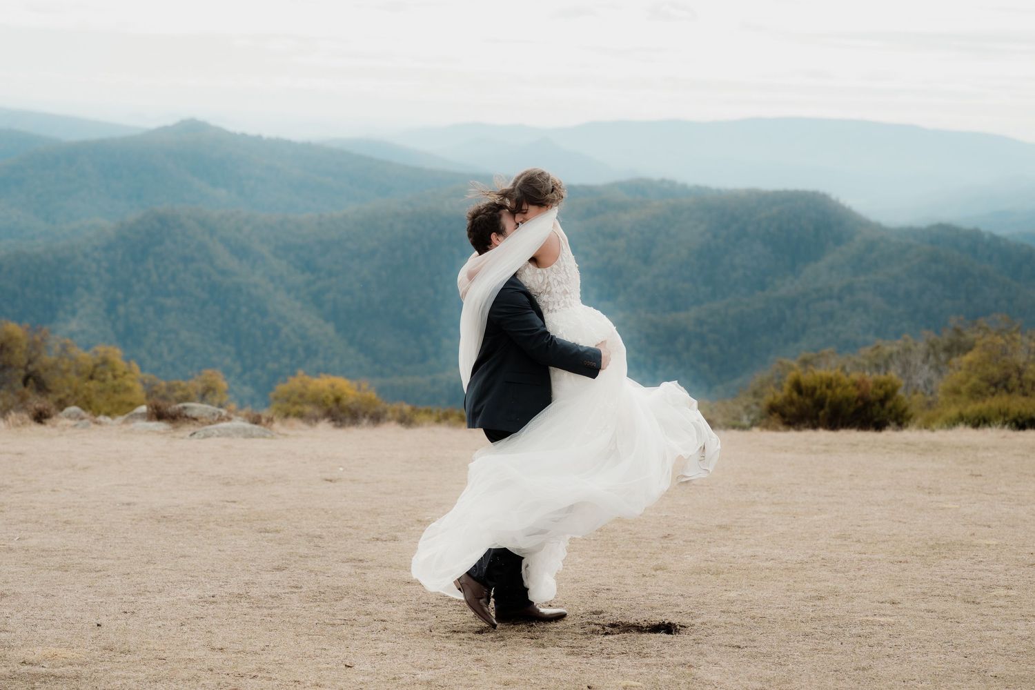 A couple in wedding attire embraces against a backdrop of misty mountain ranges during sunset at Craigs Hut