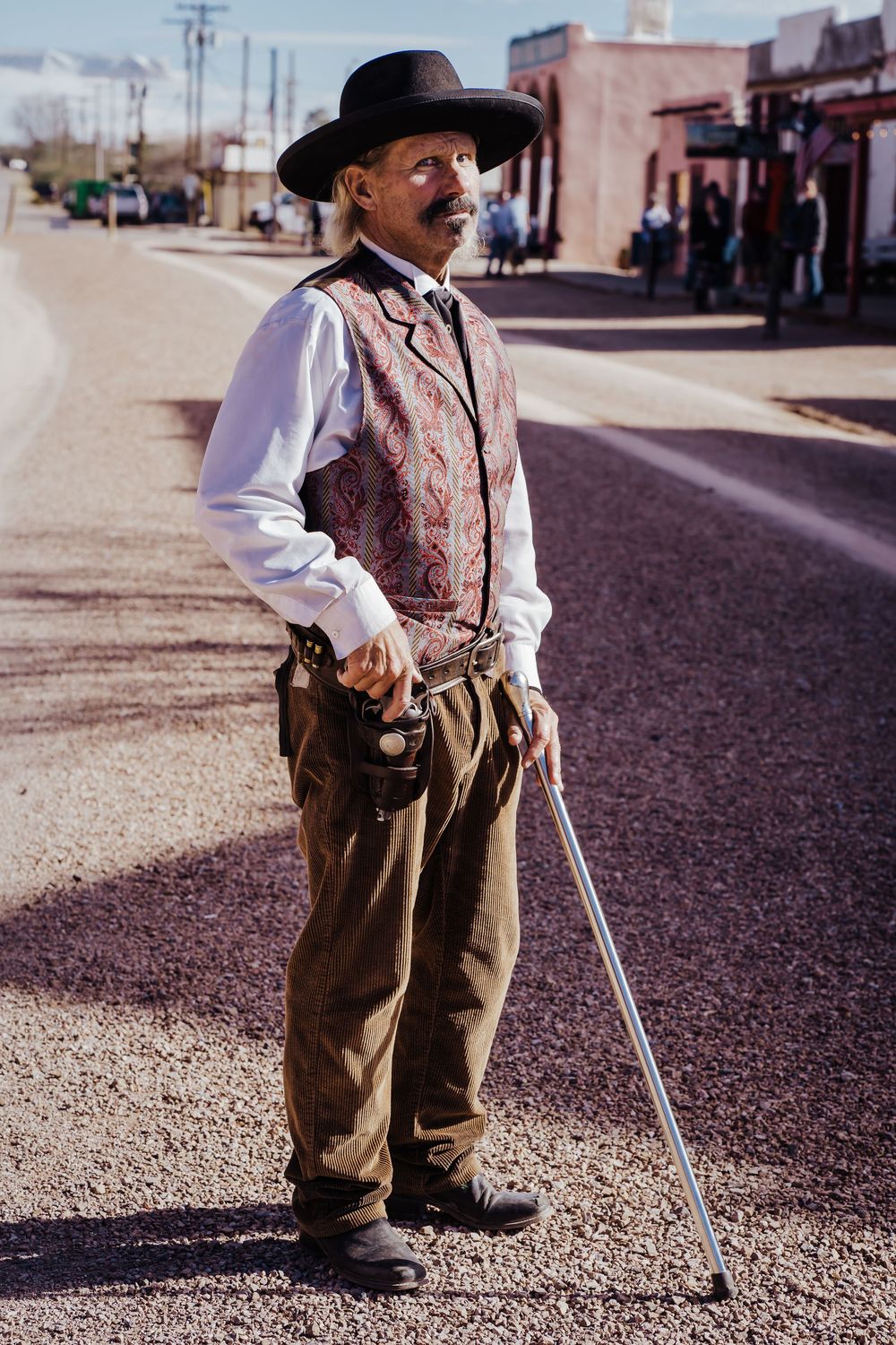Man in Old West costume with hat vest holster and cane stands on a dusty street in Tombstone.