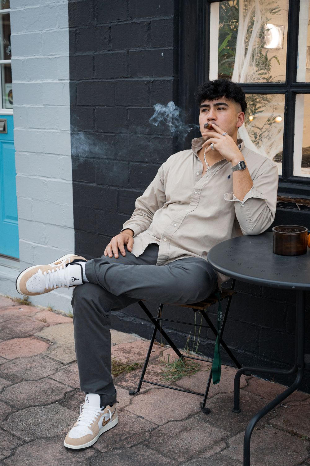 Young man in beige shirt and white sneakers smoking at an outdoor cafe table against a dark brick wall.