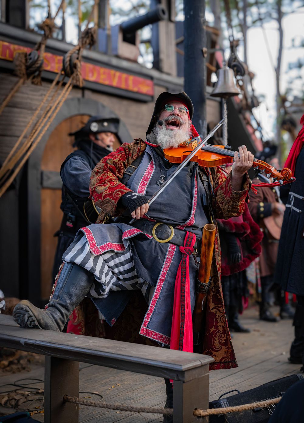 Man dressed as a pirate plays violin enthusiastically at a Renaissance or pirate festival.