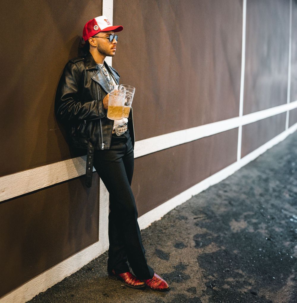 Stylish man in leather jacket red cap and flared pants leans against a wall holding a drink.