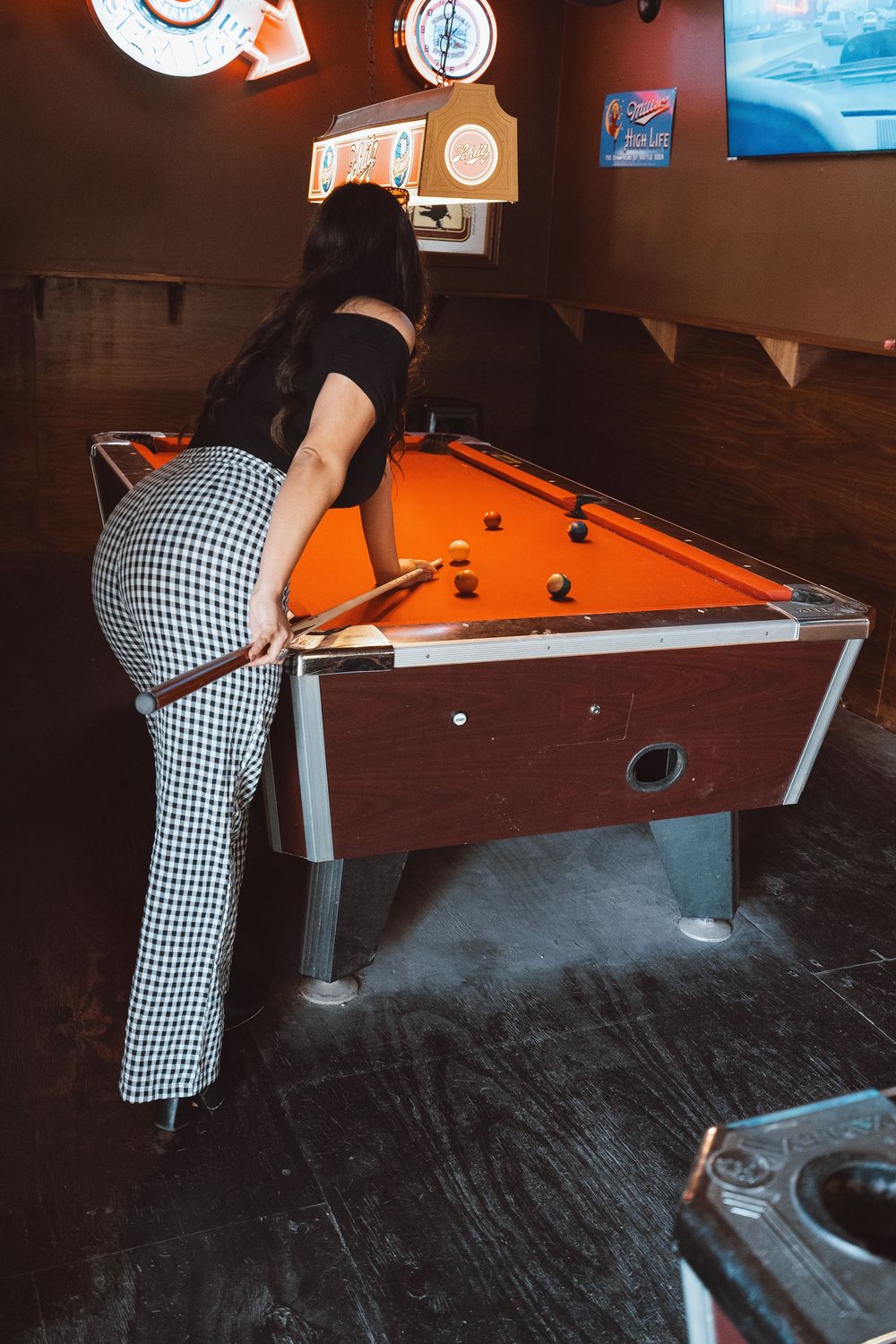 Woman in checkered pants playing pool on an orange billiard table in a dimly lit bar.