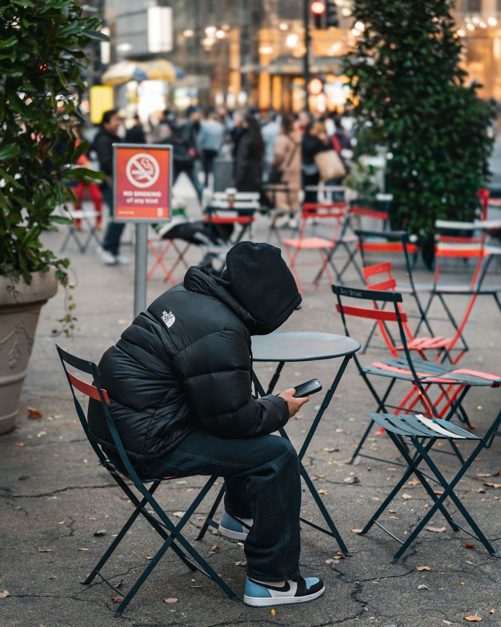 Person in black hooded puffer jacket uses smartphone at outdoor cafe table on a busy city street.