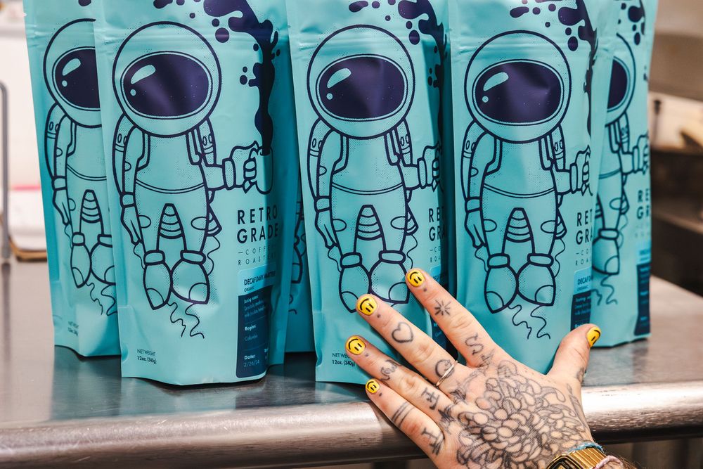 A tattooed hand reaches towards turquoise coffee bags featuring astronaut illustrations on a metal counter.