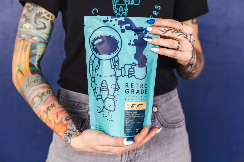 Tattooed barista holding specialty coffee bags with blue robot-themed branding designs.