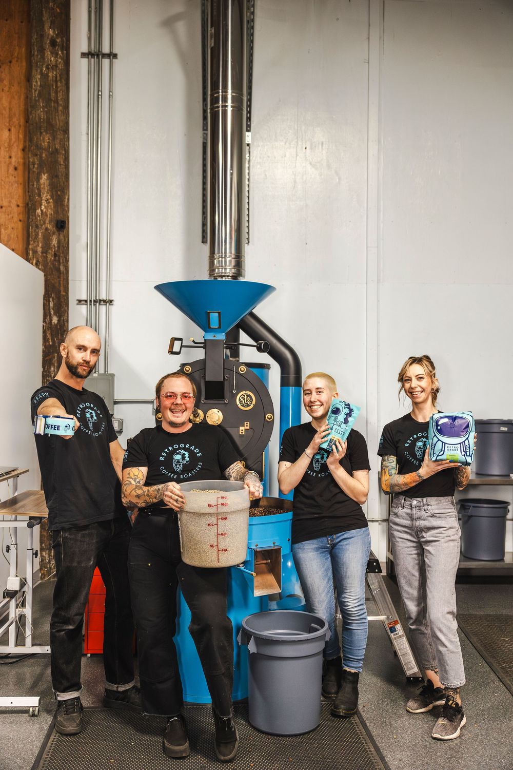Coffee roasting team poses with equipment including a blue industrial coffee roaster and storage containers in warehouse setting.