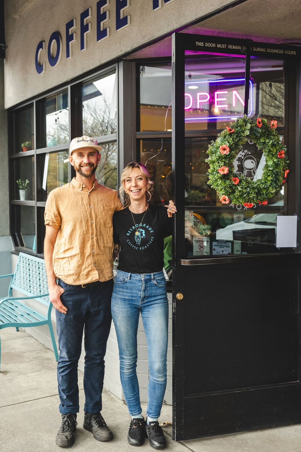Two people stand together outside a coffee shop entrance with holiday wreaths and decorations visible in the background.