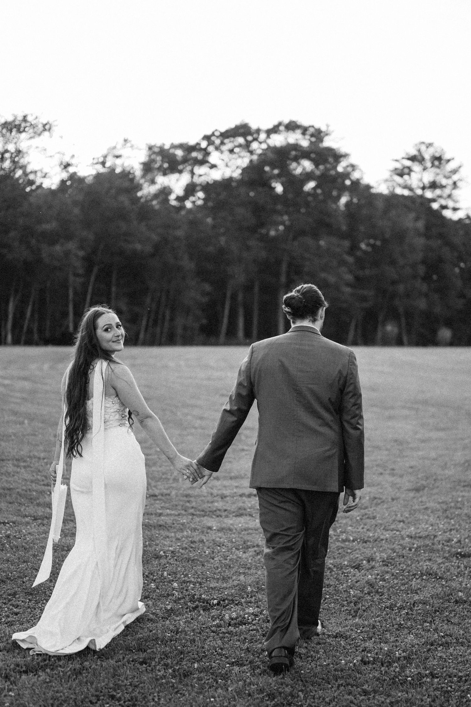 A bride and groom walk hand in hand through a grassy field with pine trees in the background.
