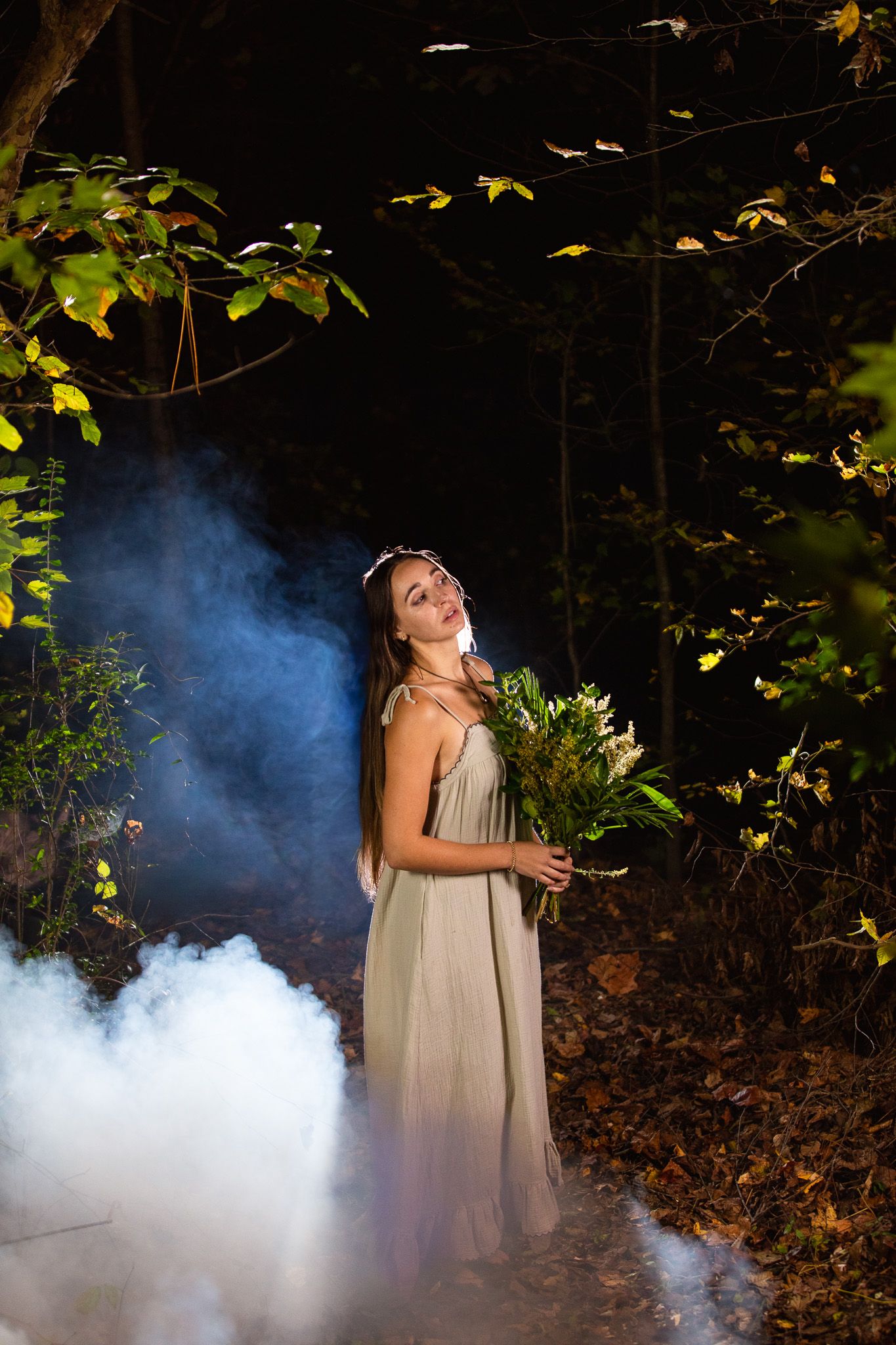 Atmospheric photo showing a woman emerging through white smoke effects in a dark forest setting holding flowers