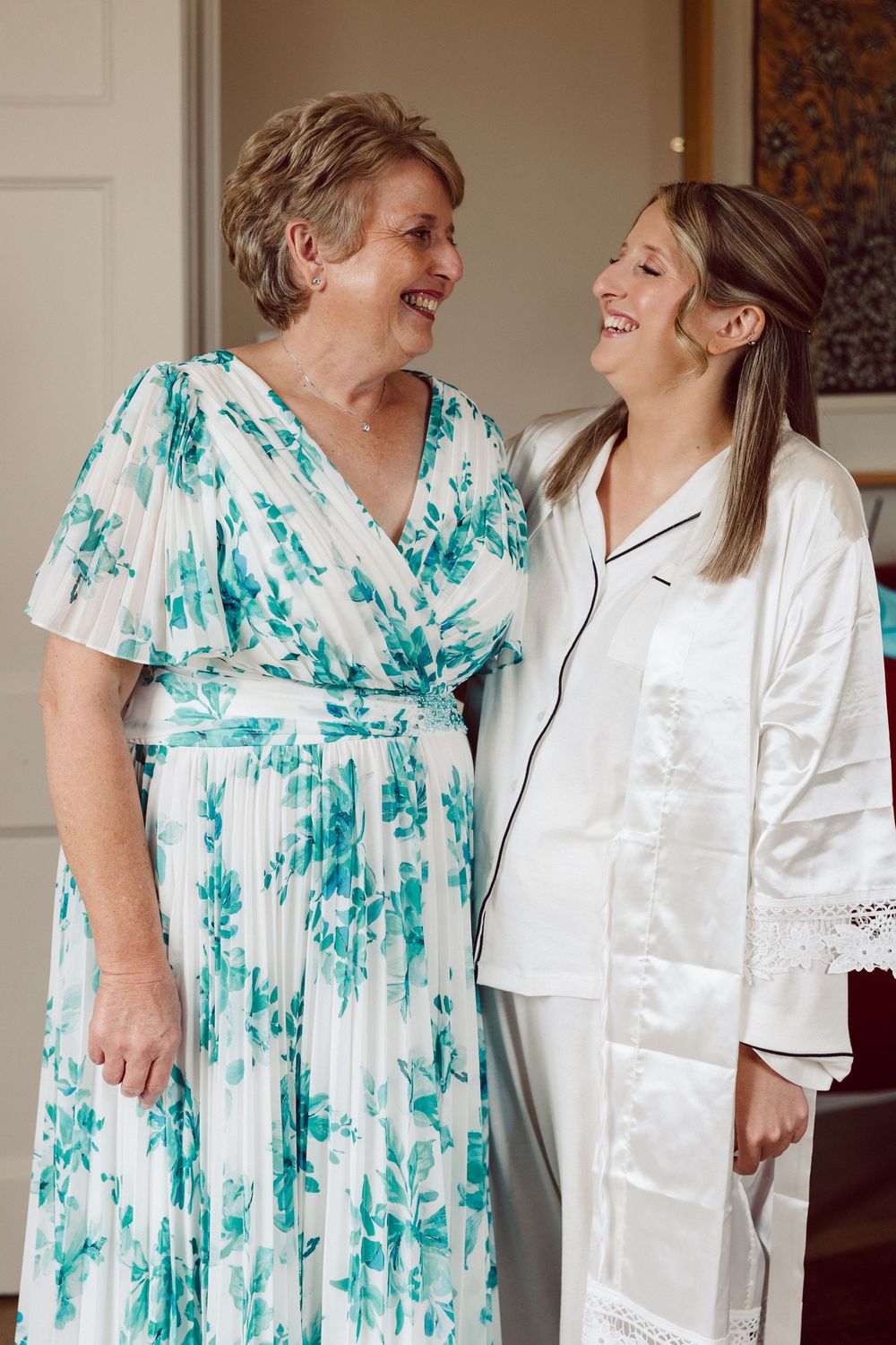 Two people share a joyful laugh together, one wearing a floral turquoise dress and the other in white sleepwear.