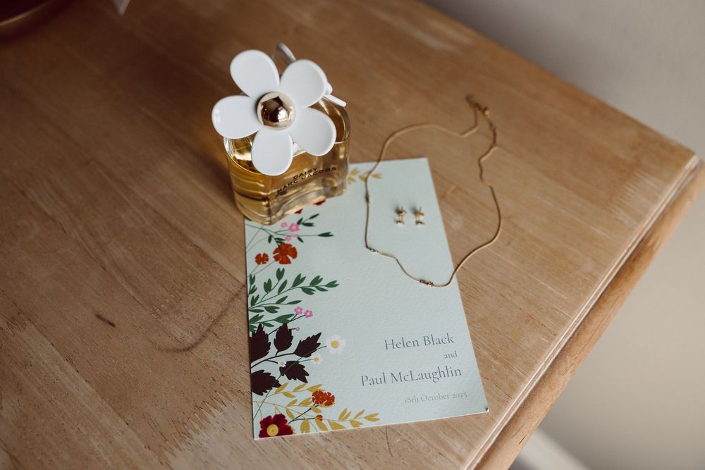 A wedding invitation card with floral design lies on a wooden table next to a white paper flower decoration.