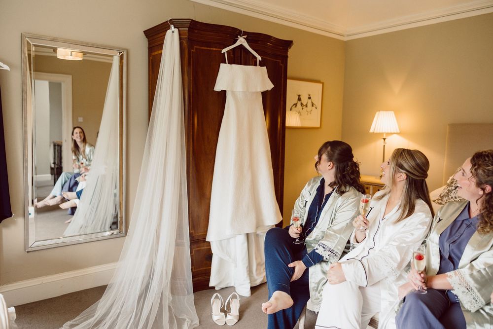 A white wedding dress hangs in a wardrobe while people in robes sit on a couch in a bridal suite.