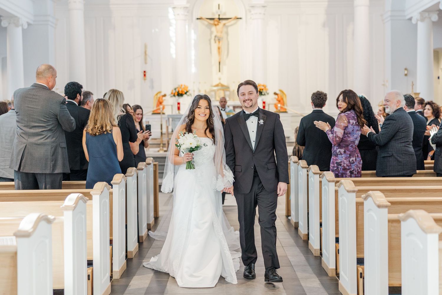 A wedding ceremony taking place in a traditional white church with guests seated in wooden pews.