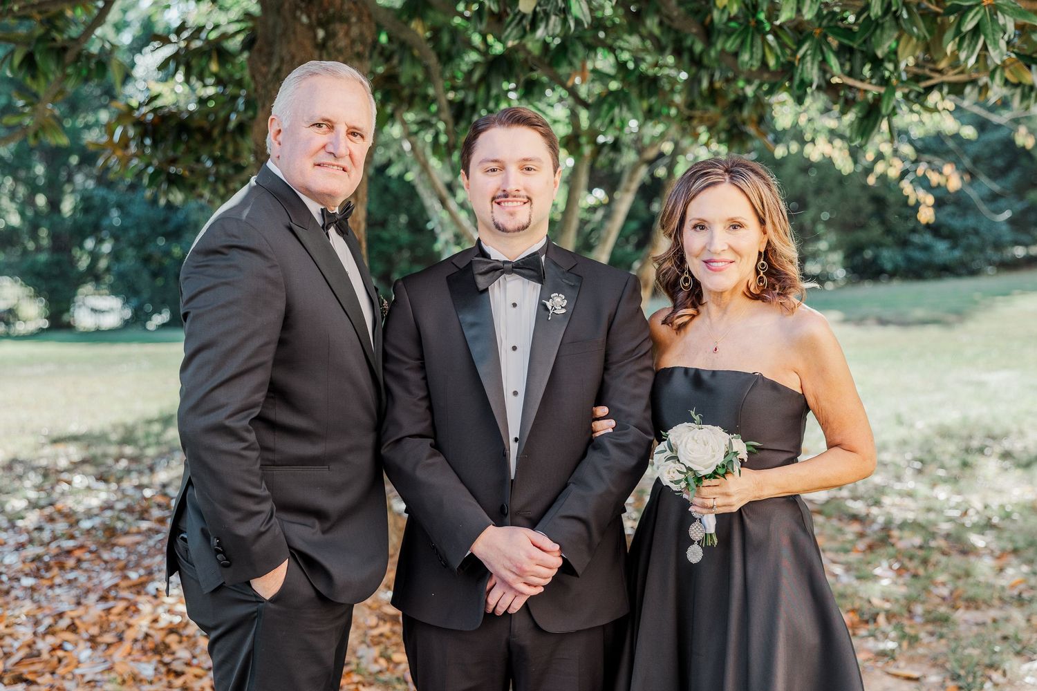 A wedding party poses together outdoors in formal attire during an autumn day with fallen leaves on the ground.