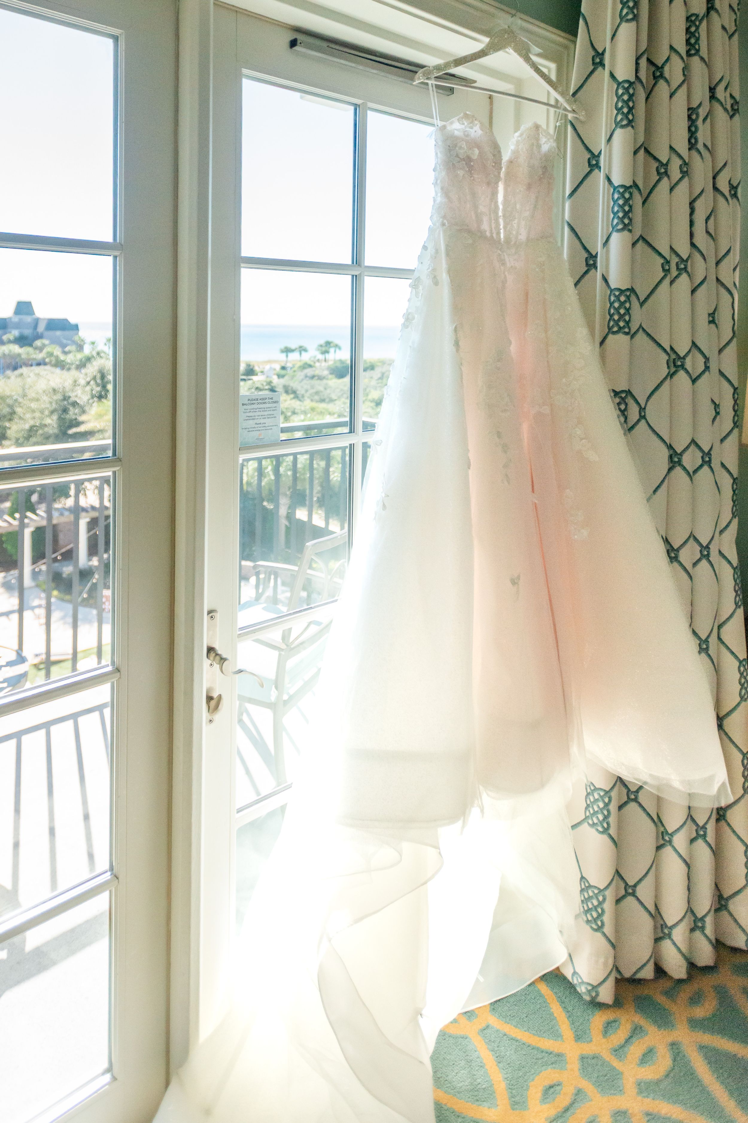 White wedding dress hanging by sunlit French doors with patterned curtains and ocean view.