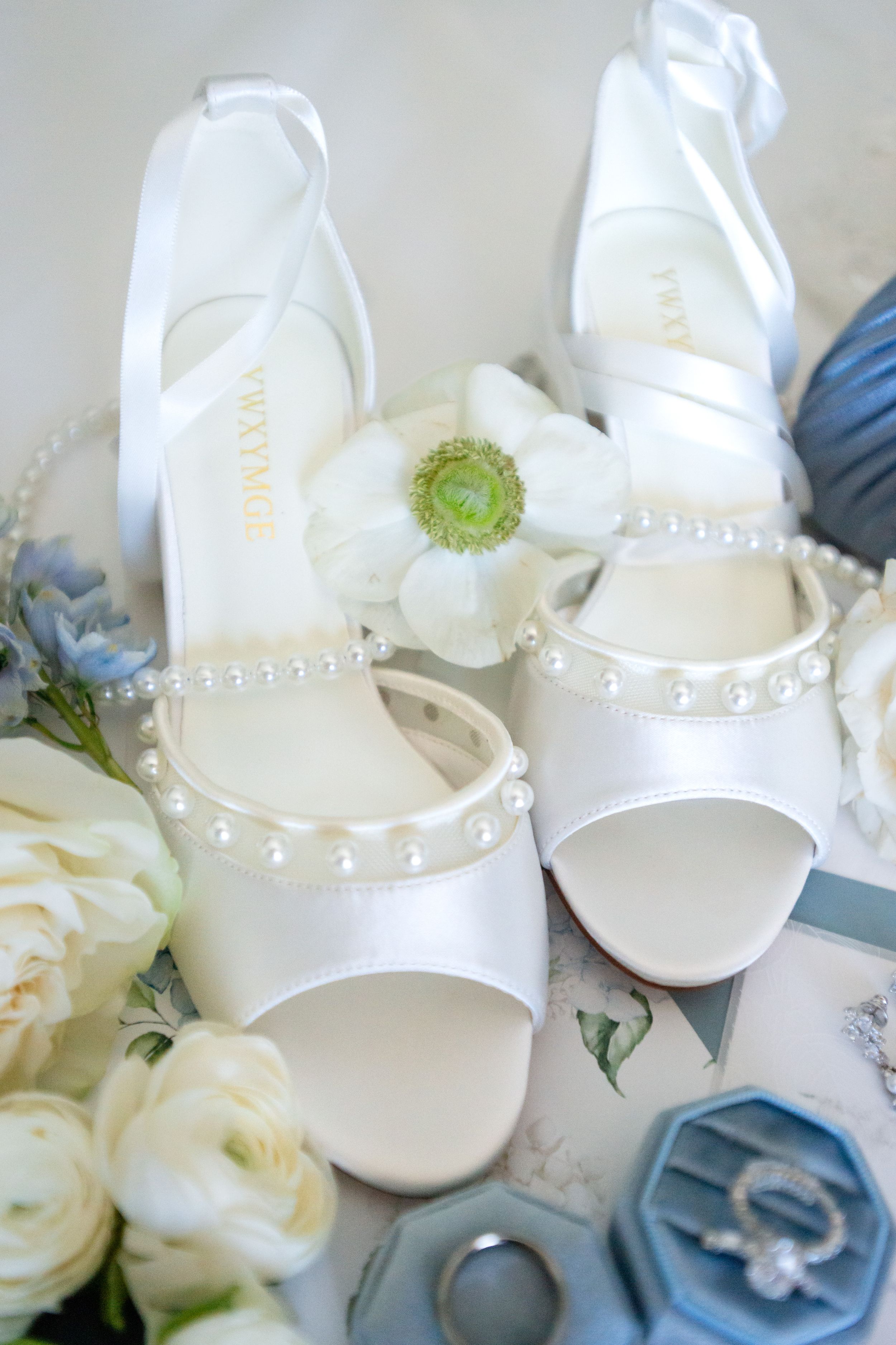 Elegant white bridal shoes with pearl details displayed alongside white roses and blue floral accents.