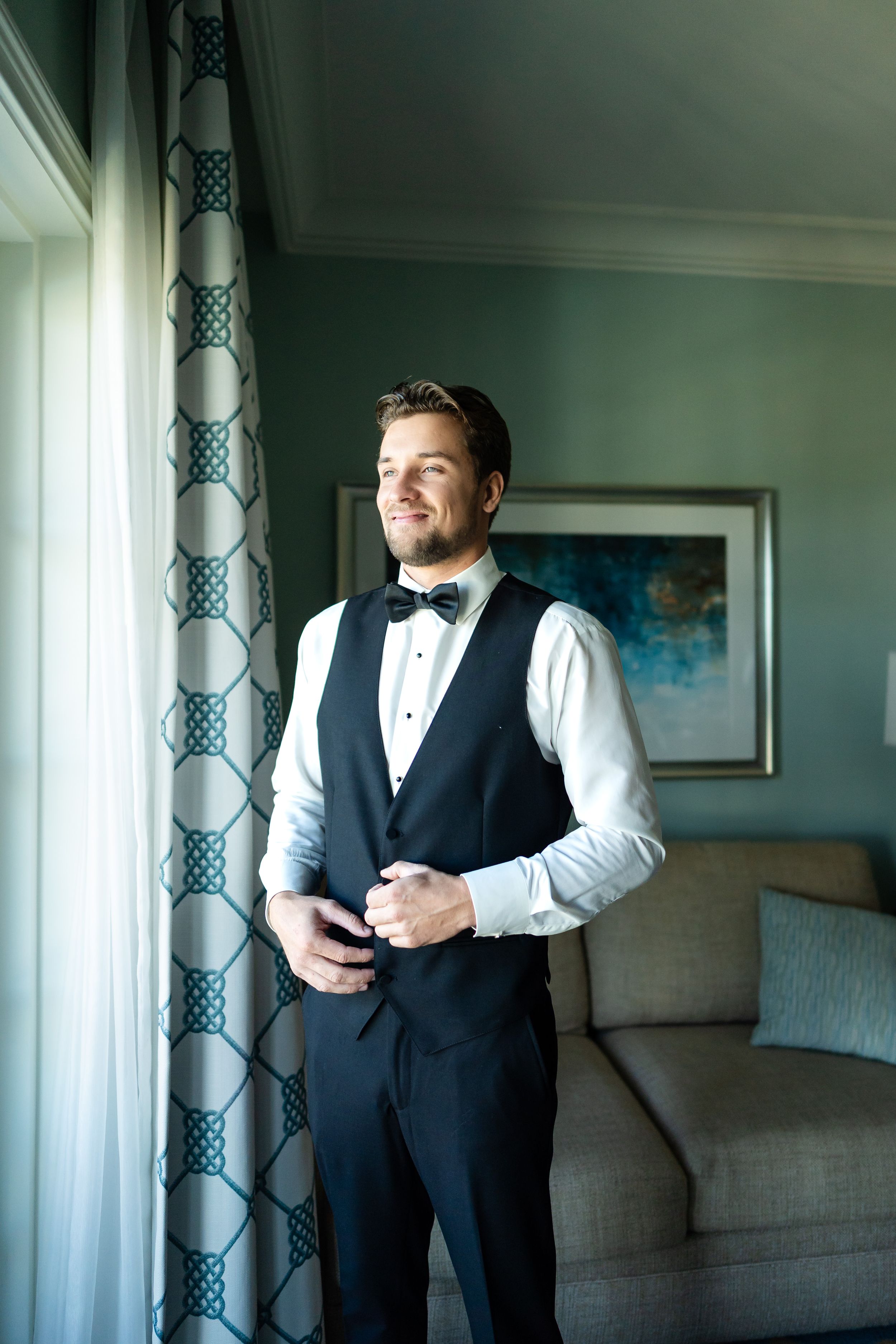 Groom in formal attire adjusts his bowtie while getting ready in a sunlit room.