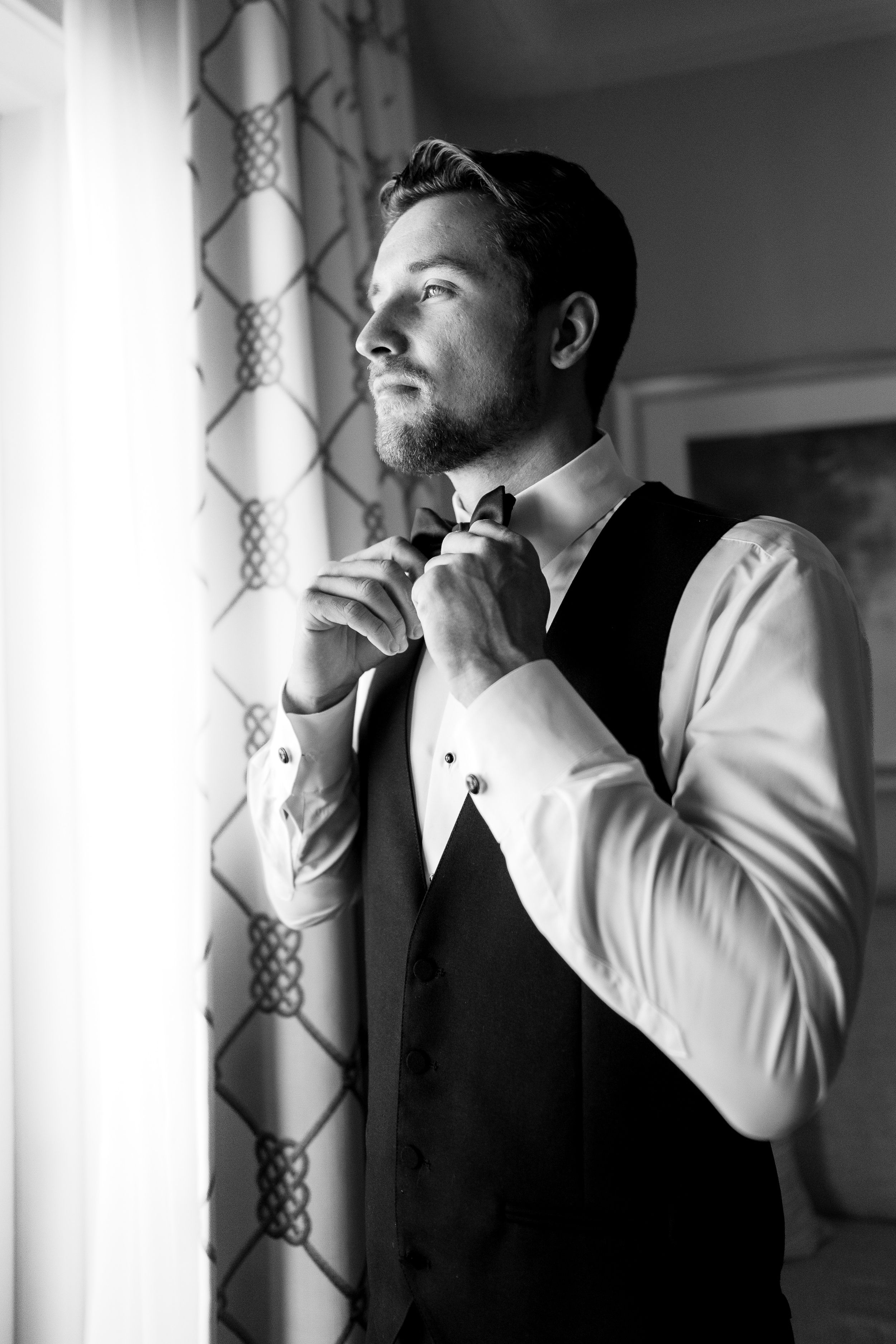 Black and white portrait of a groom adjusting his bowtie while getting ready.