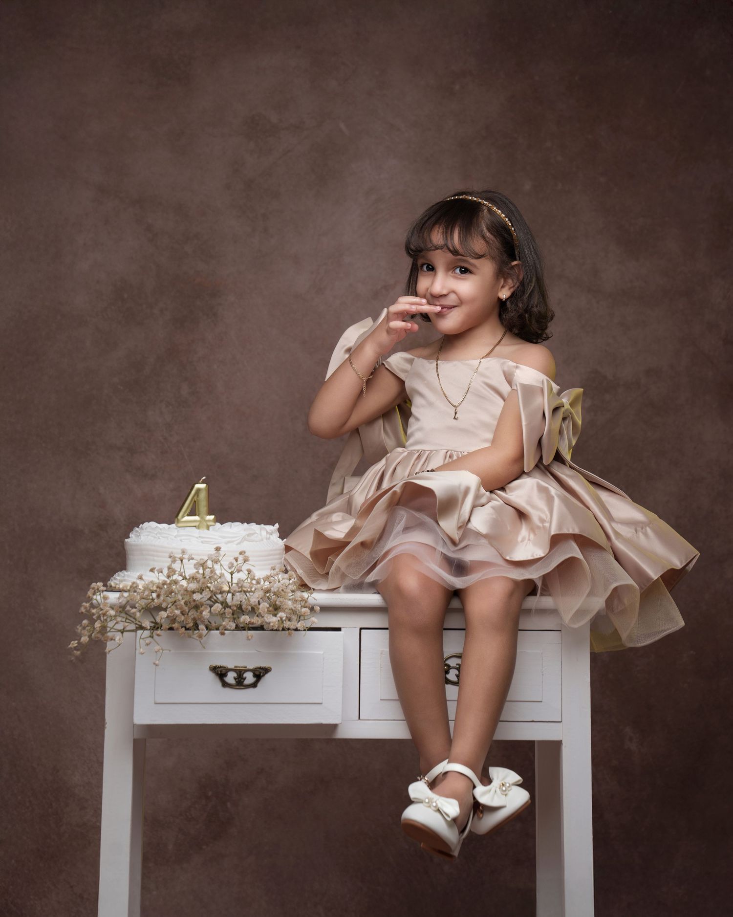 A child in a pink dress sits playfully on a white bench with delicate white flowers in a rustic studio setting.