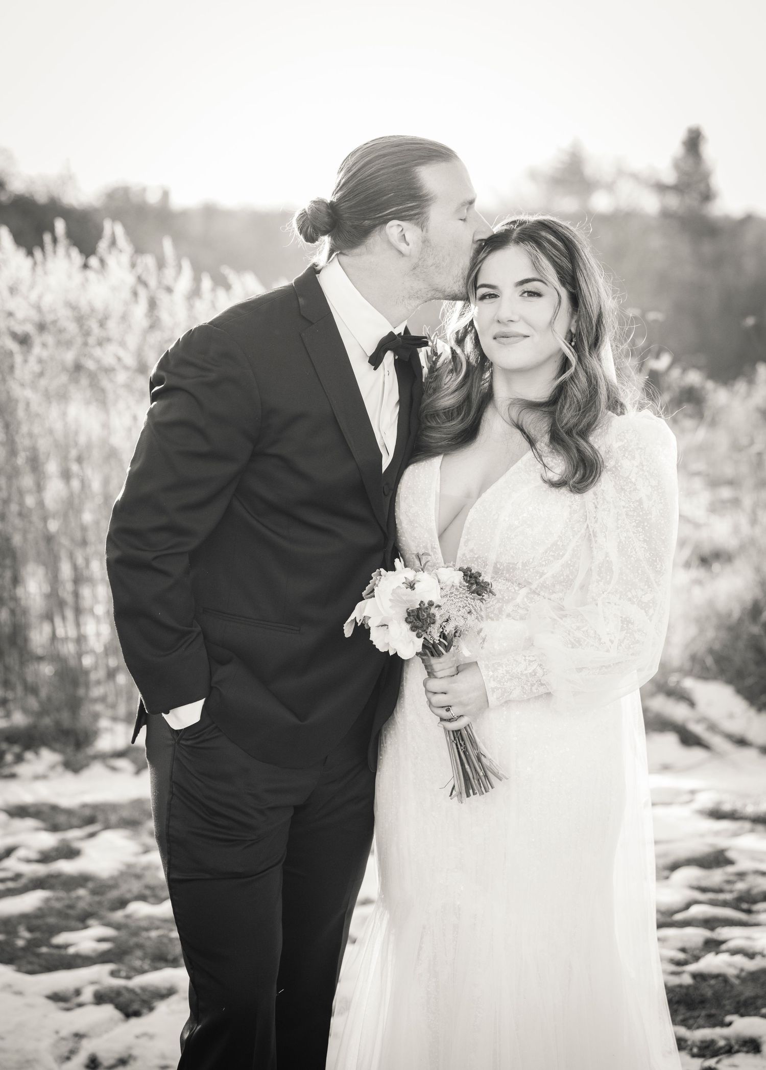 Black and white outdoor wedding photo of newlyweds in formal attire with white bouquet amid tall grass in natural setting.
