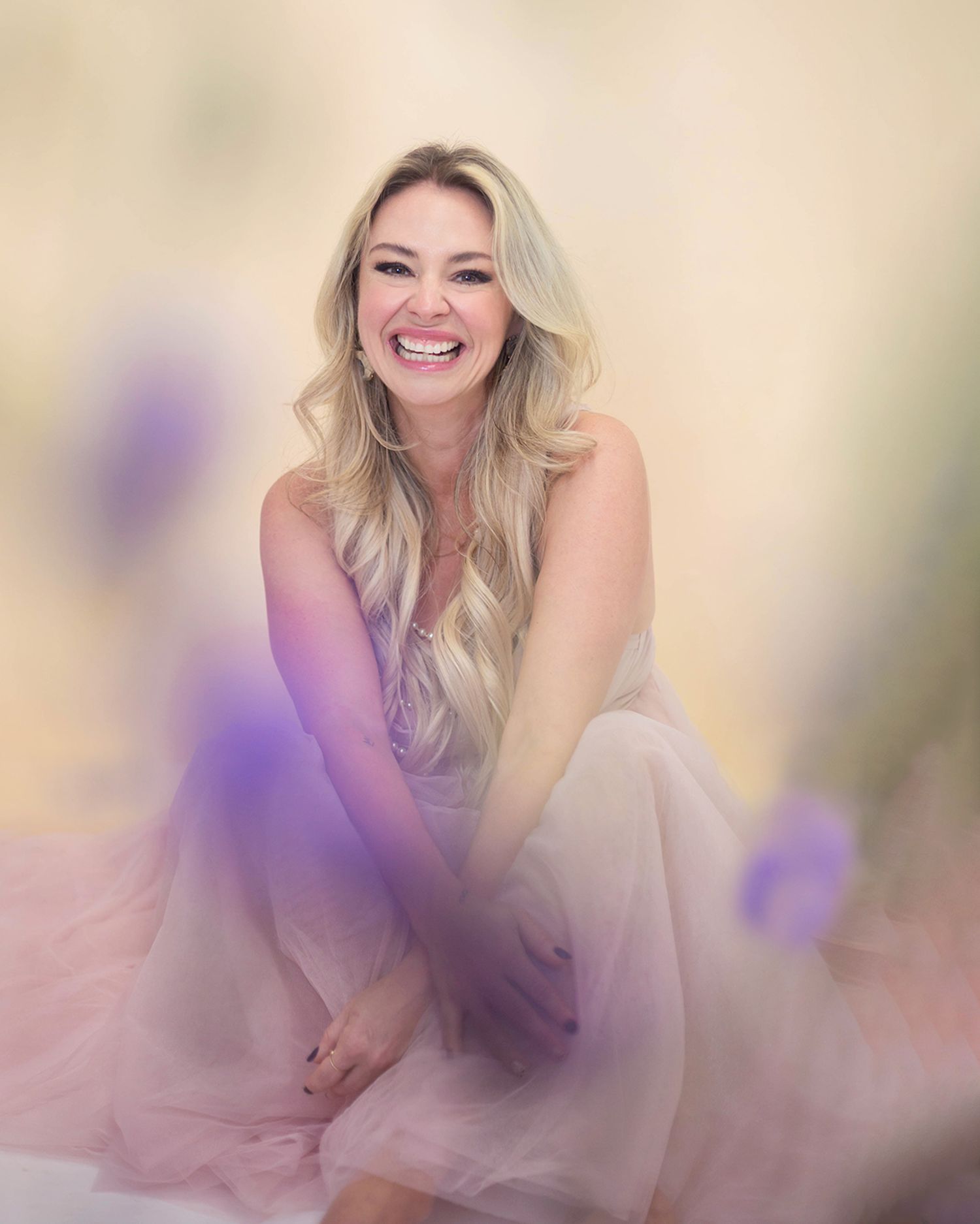 A woman in soft pink lighting poses joyfully with a natural smile and blonde hair against a dreamy pastel background.