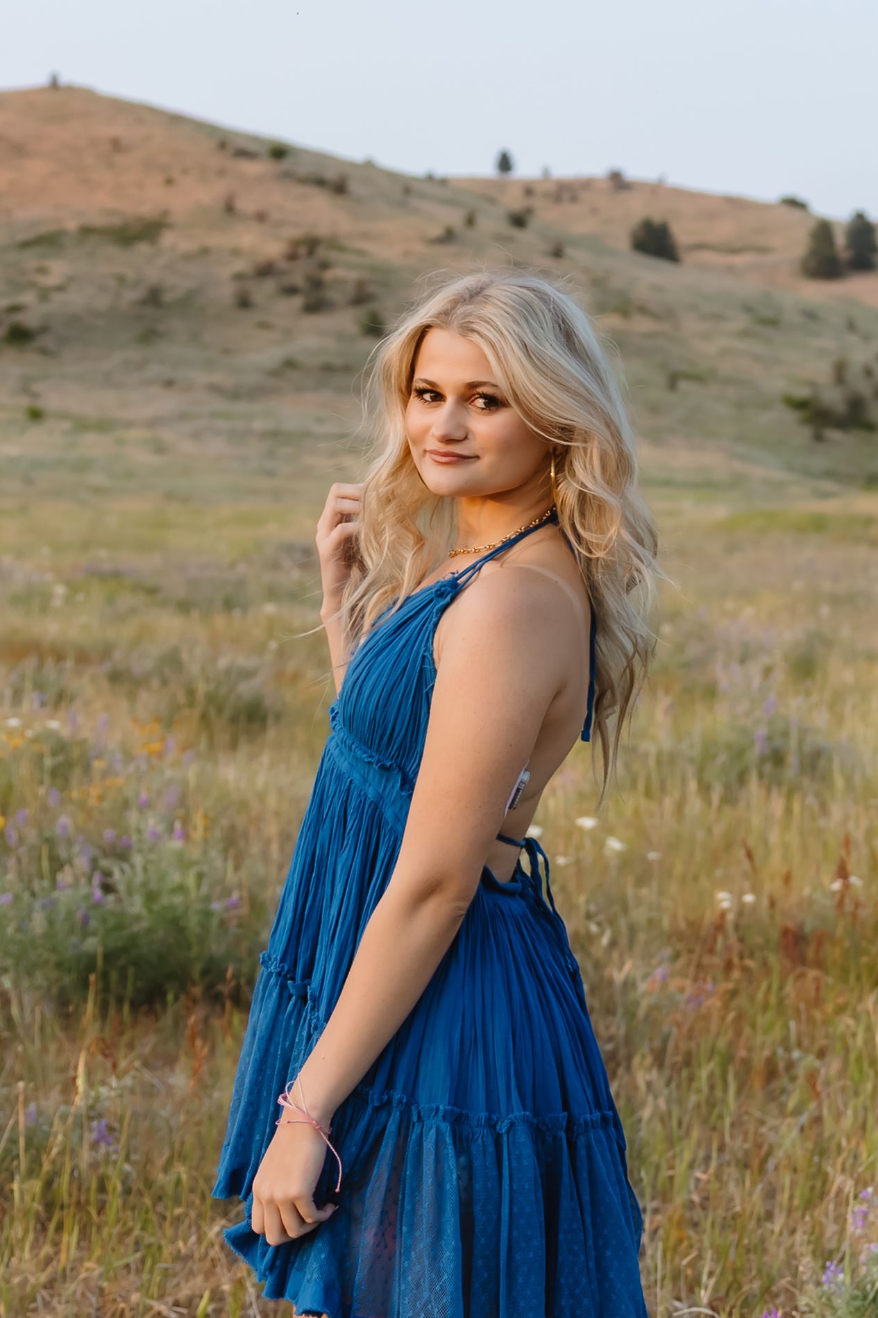 A person in a flowing royal blue dress poses in a grassy meadow with rolling hills in the background during golden hour.