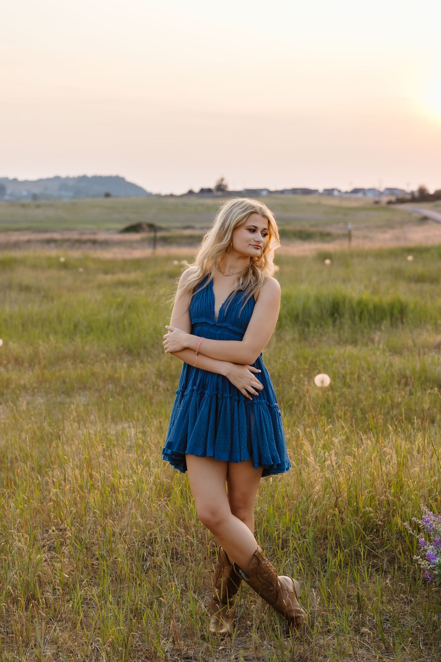 A woman in a navy blue dress and cowboy boots stands in a golden field at sunset.