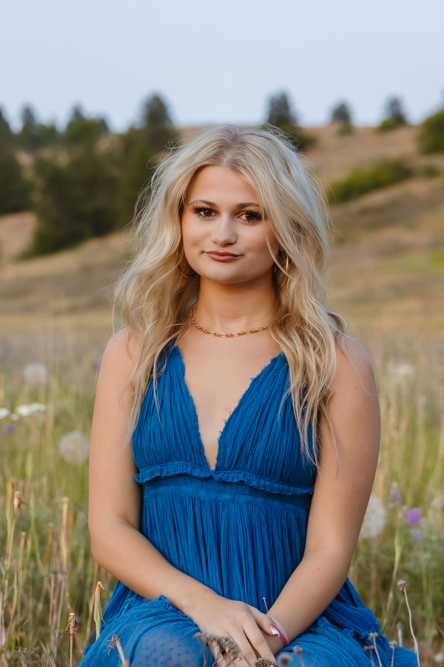 A person in a flowing blue dress poses outdoors in a field with wildflowers during golden hour.