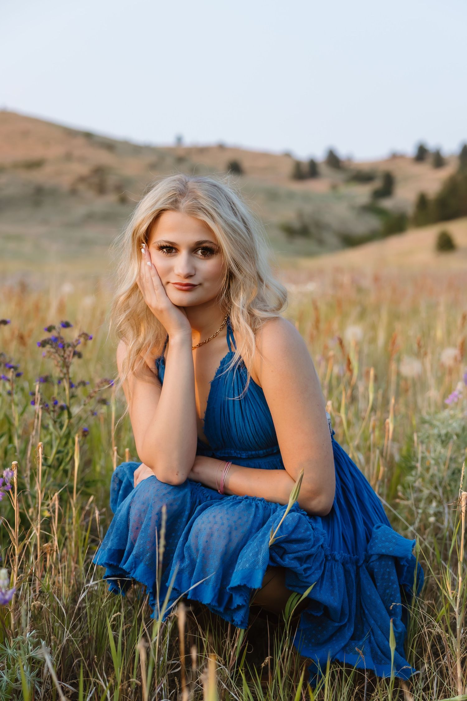 A person in a blue dress sits among wildflowers in a rolling meadow during golden hour.
