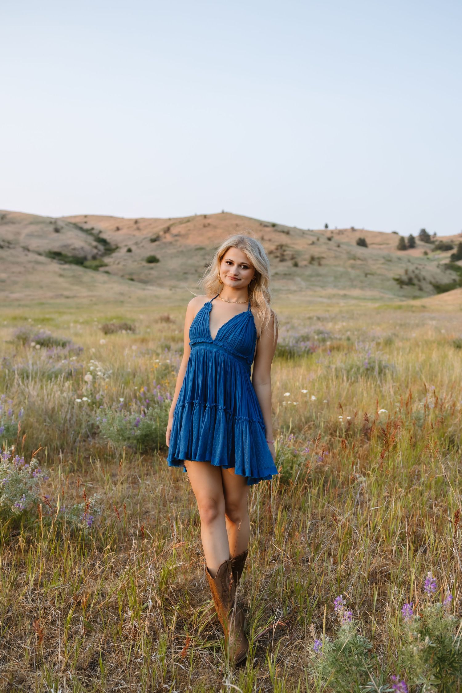 Woman wearing a flowy blue dress stands in a grassy field with mountains in the background at sunset.