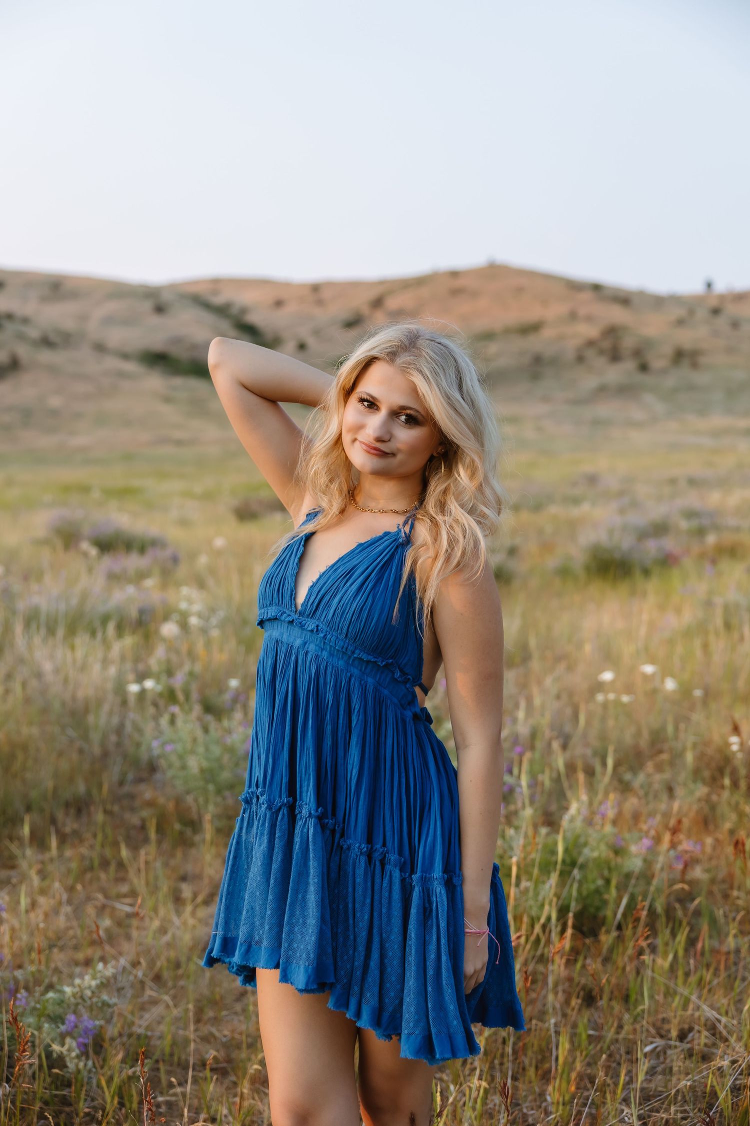 A woman in a flowing blue sundress poses in a summer field with rolling hills in the background.