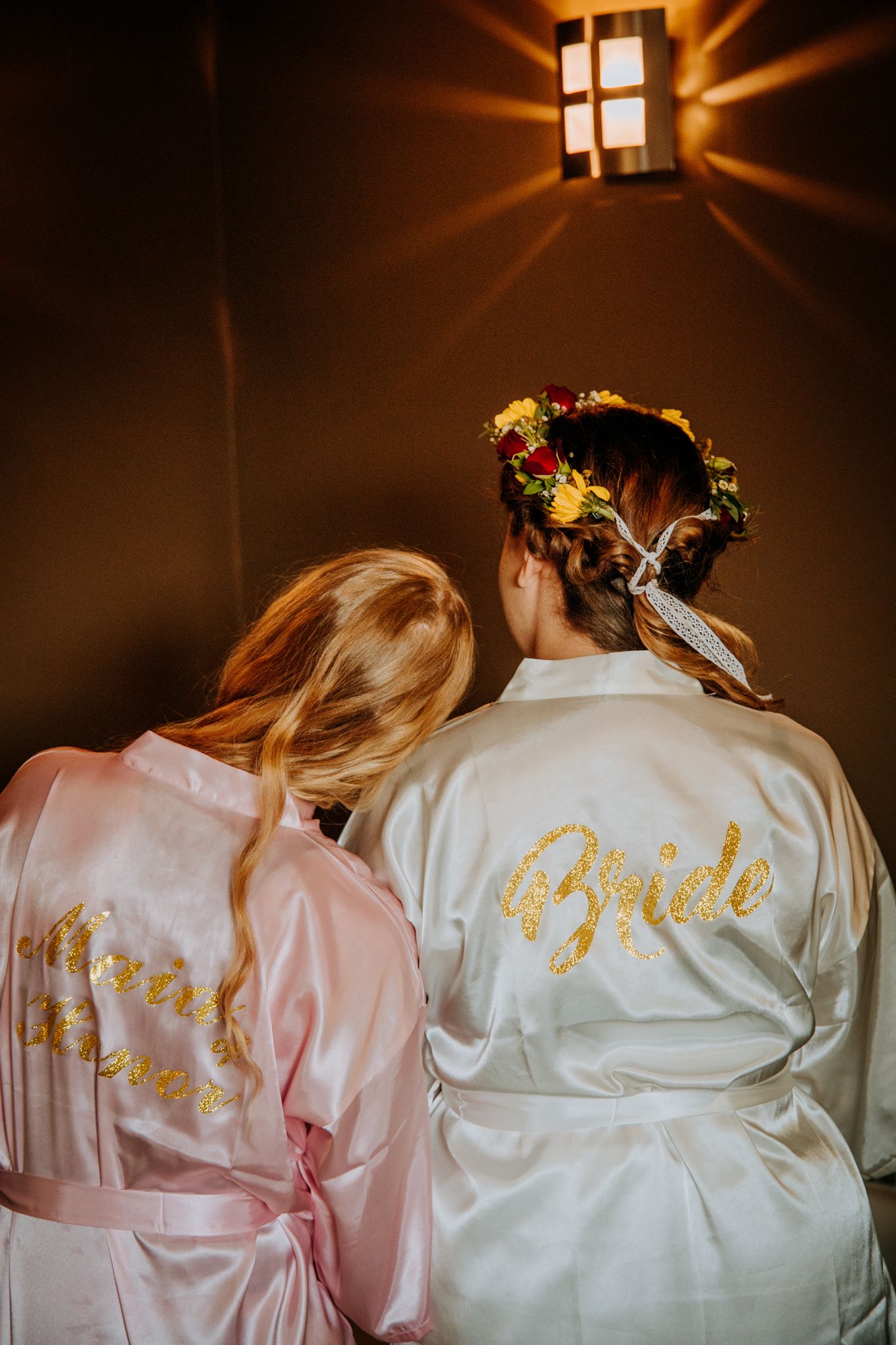 Bridal party wearing matching silk robes in pink and white with gold lettering shown from behind in a dark room.