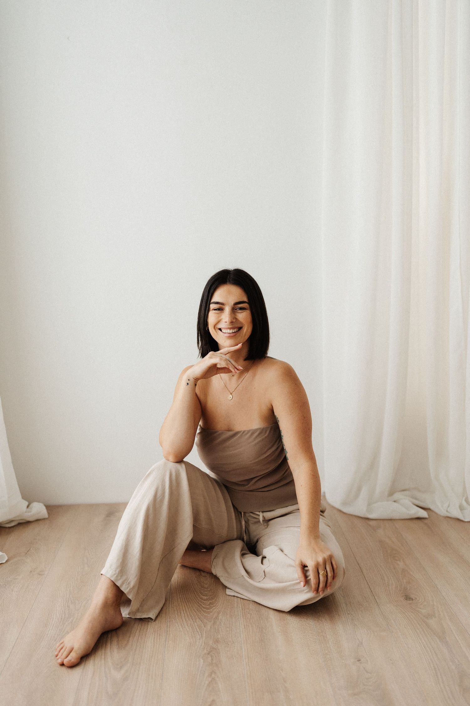 A woman in a beige dress poses naturally on a wooden floor against a white curtain backdrop in a photo series.