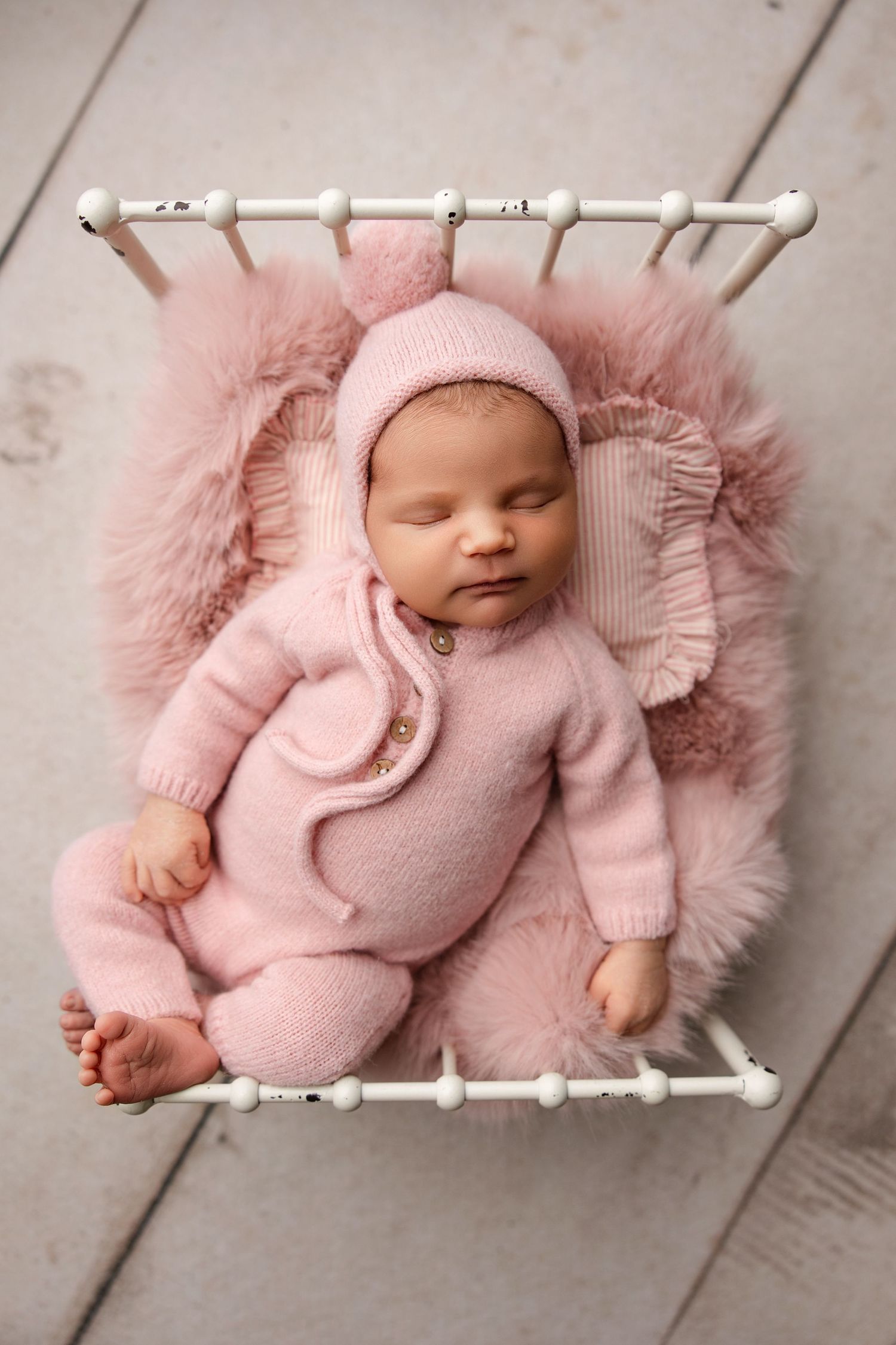 Sleeping baby wearing soft pink knit outfit lying on fluffy pink blanket in small white decorative bed.