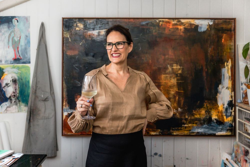 Artist stands in front of abstract painting in studio wearing beige top and black skirt.