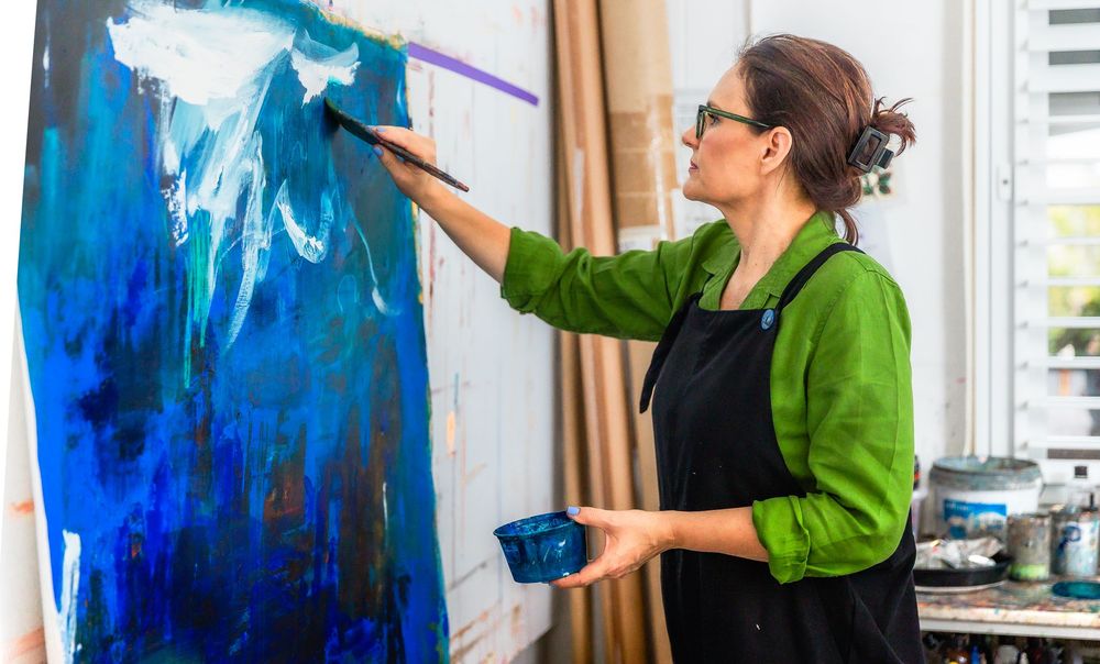 Artist in green shirt working on vibrant blue abstract painting across multiple canvas panels in bright studio space.