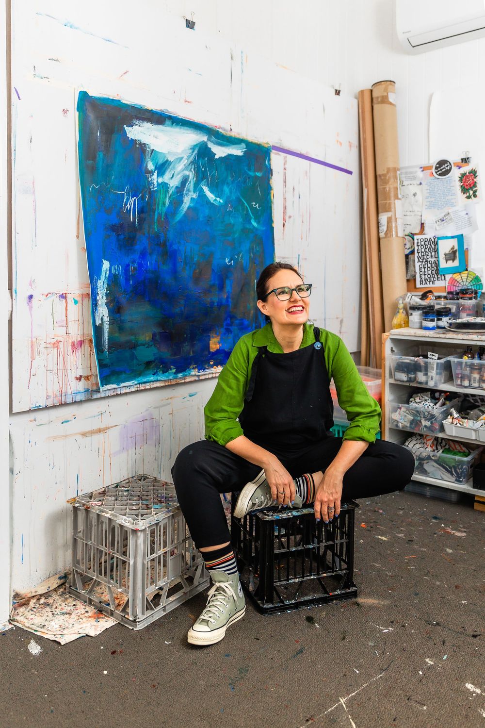 Artist in green sweater and black apron sits in studio with vibrant blue abstract painting in background.