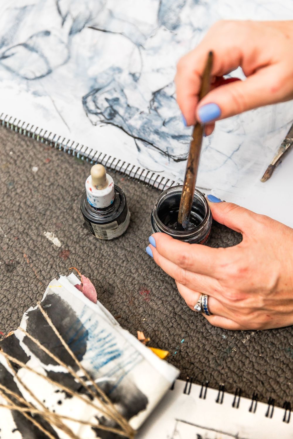 Hands dipping brushes into black ink or paint on a spiral-bound sketchbook.