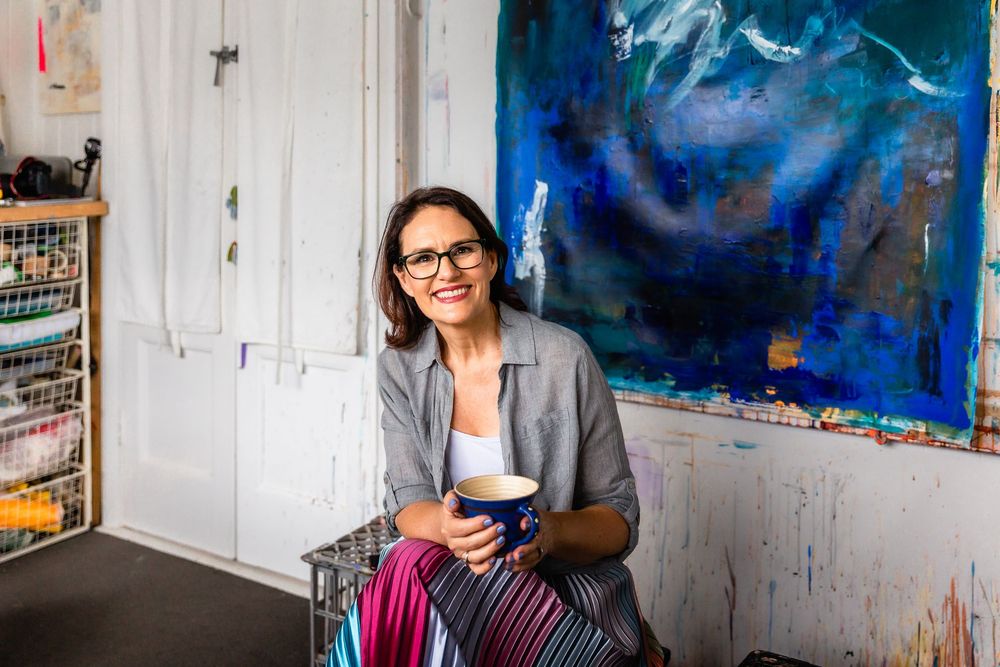 Artist in gray blazer and glasses sits smiling in studio with vibrant blue abstract painting in background.