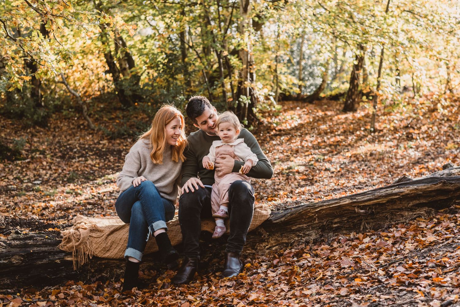 A family of three sitting on a tree trunk in an autumnal woodland setting.