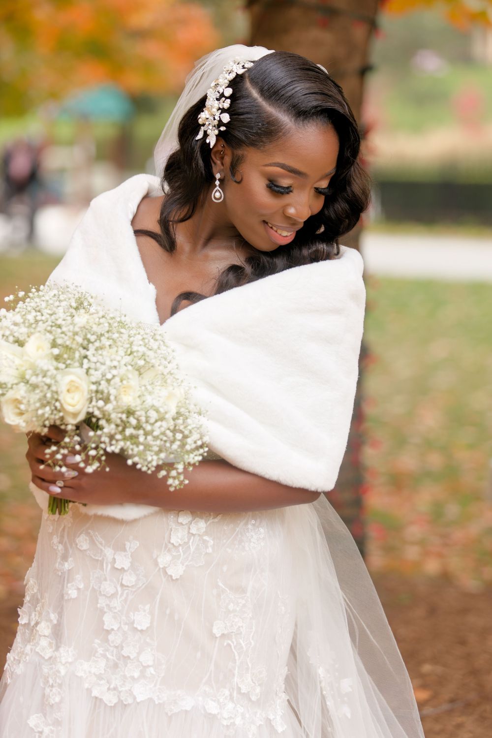 Elegant Black bride in lace wedding gown and white fur wrap gazing at bouquet of white roses amid fall foliage.