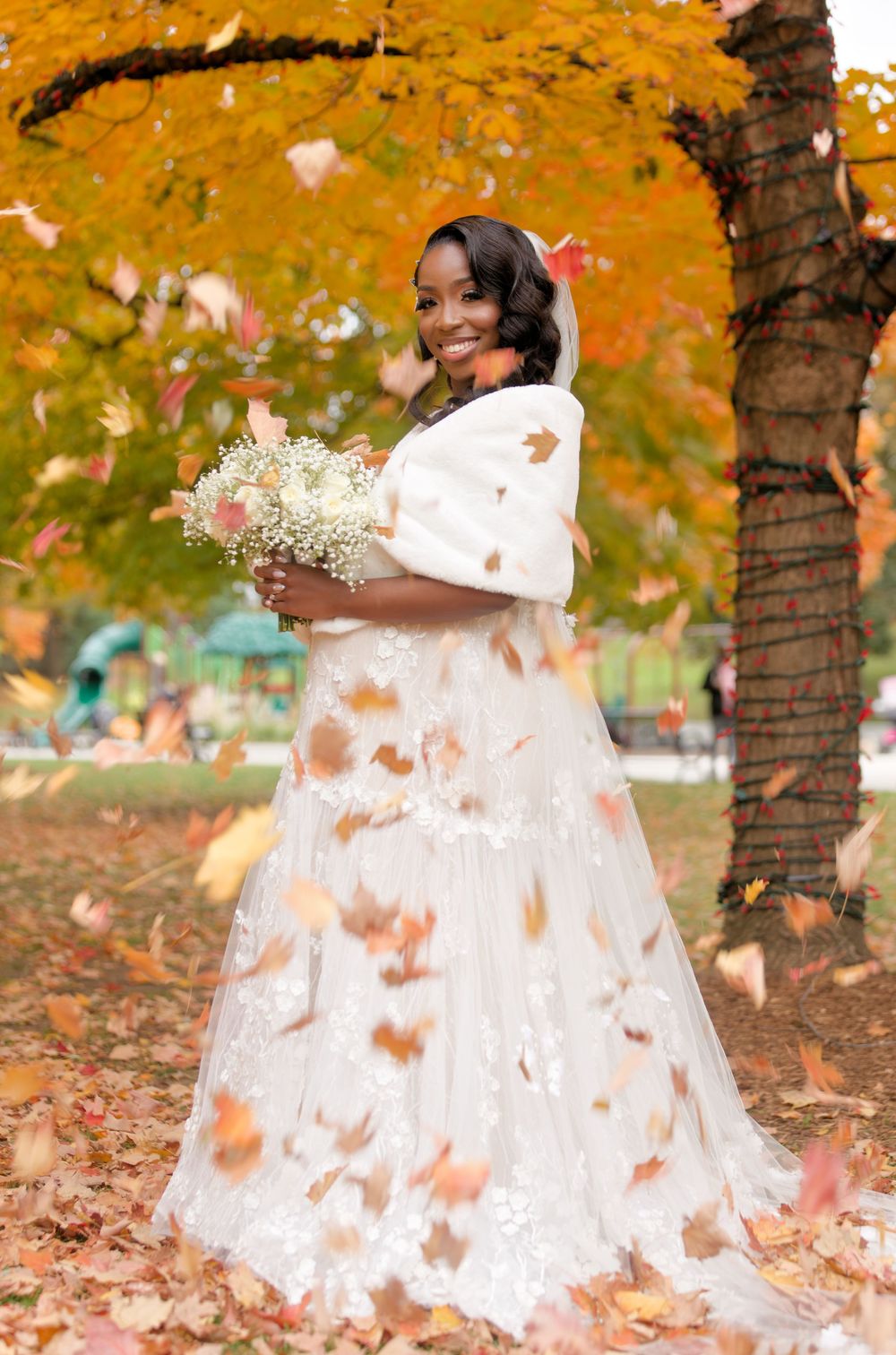 Radiant Black bride in white lace gown and fur wrap holding bouquet, surrounded by falling autumn leaves in golden park.