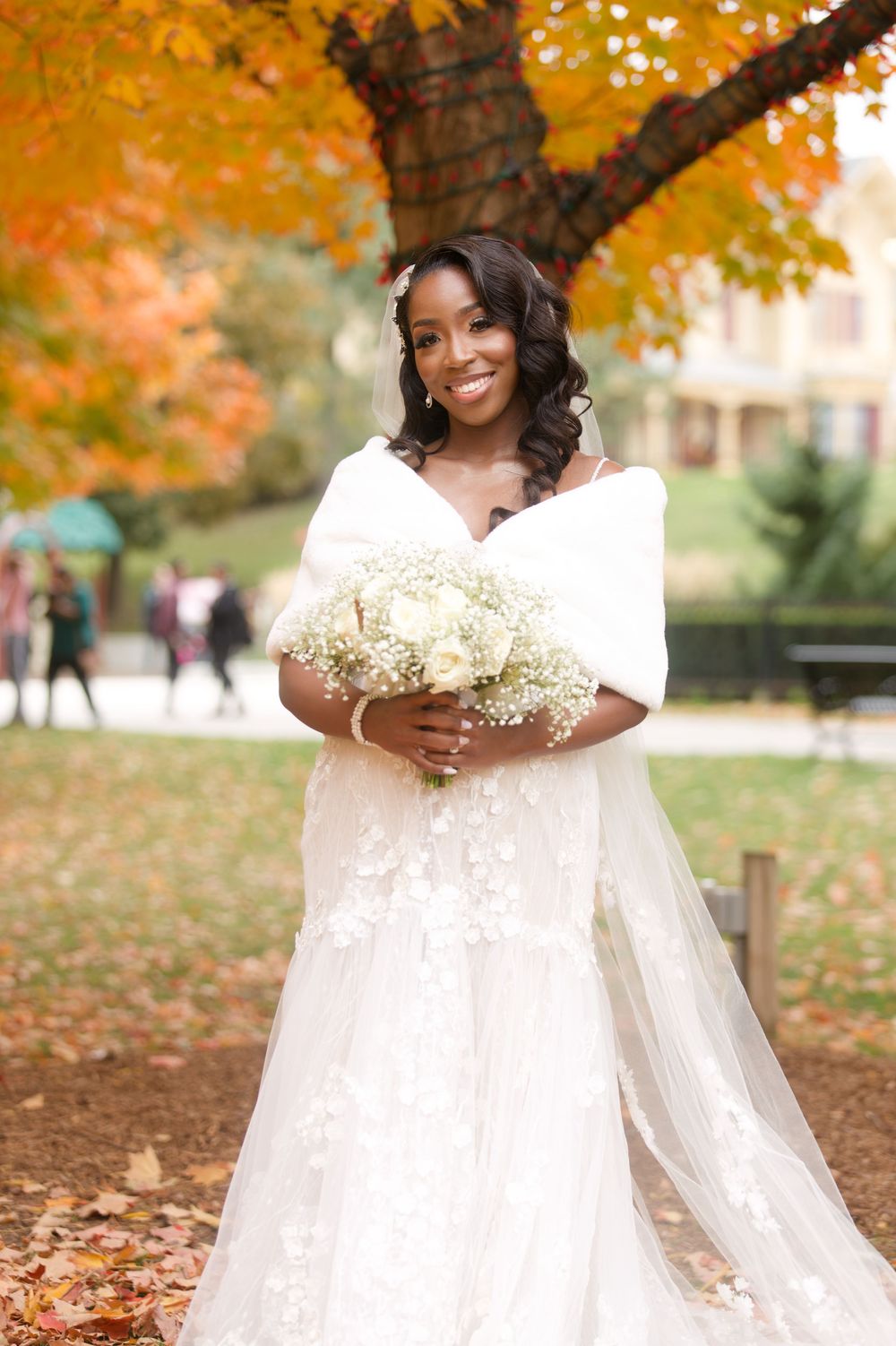 Beautiful Black bride smiling in white lace gown with fur stole, holding white bouquet before vibrant orange autumn tree.