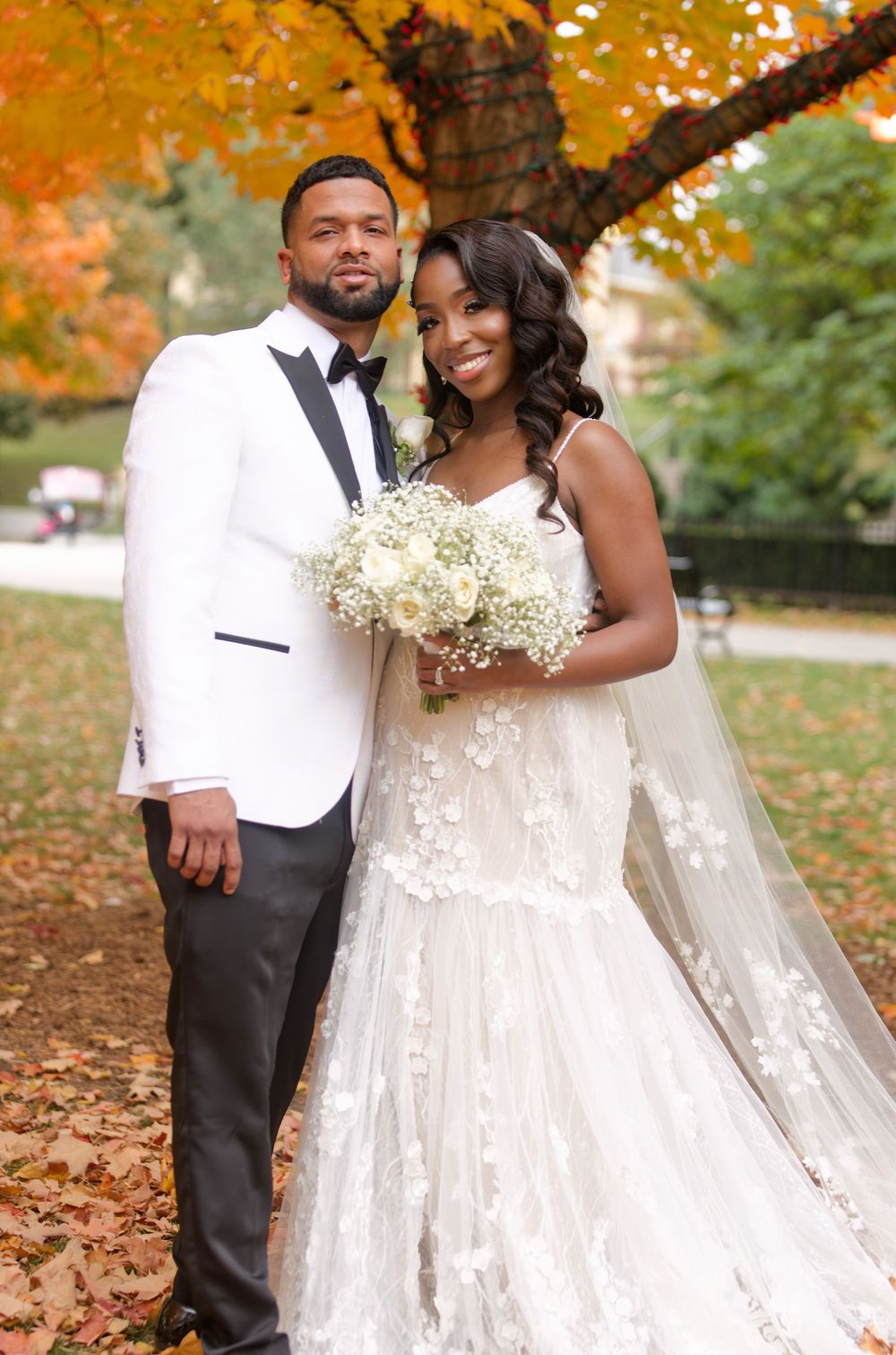 A bride and groom pose together under a golden autumn tree holding white flowers.